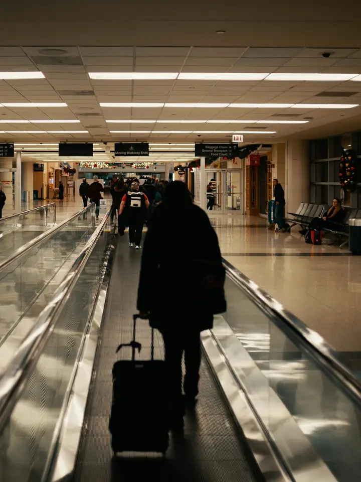 A traveler pulls a rolling carry-on down an airport moving walkway, with other travelers and their backpacks up ahead.